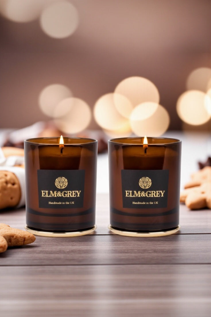 Two Elm & Grey candles on a wooden surface with blurred lights in the background