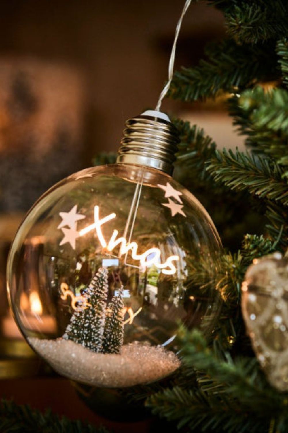 Decorative light bulb ornament with 'Xmas' and Christmas tree inside, hanging on a tree.