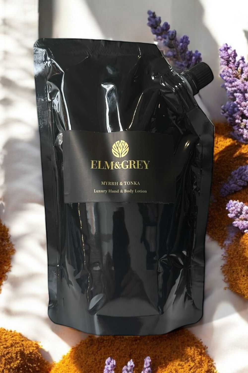 Black packaging of Elm & Grey myrrh & tonka luxury hand & body lotion on a textured surface with flowers.