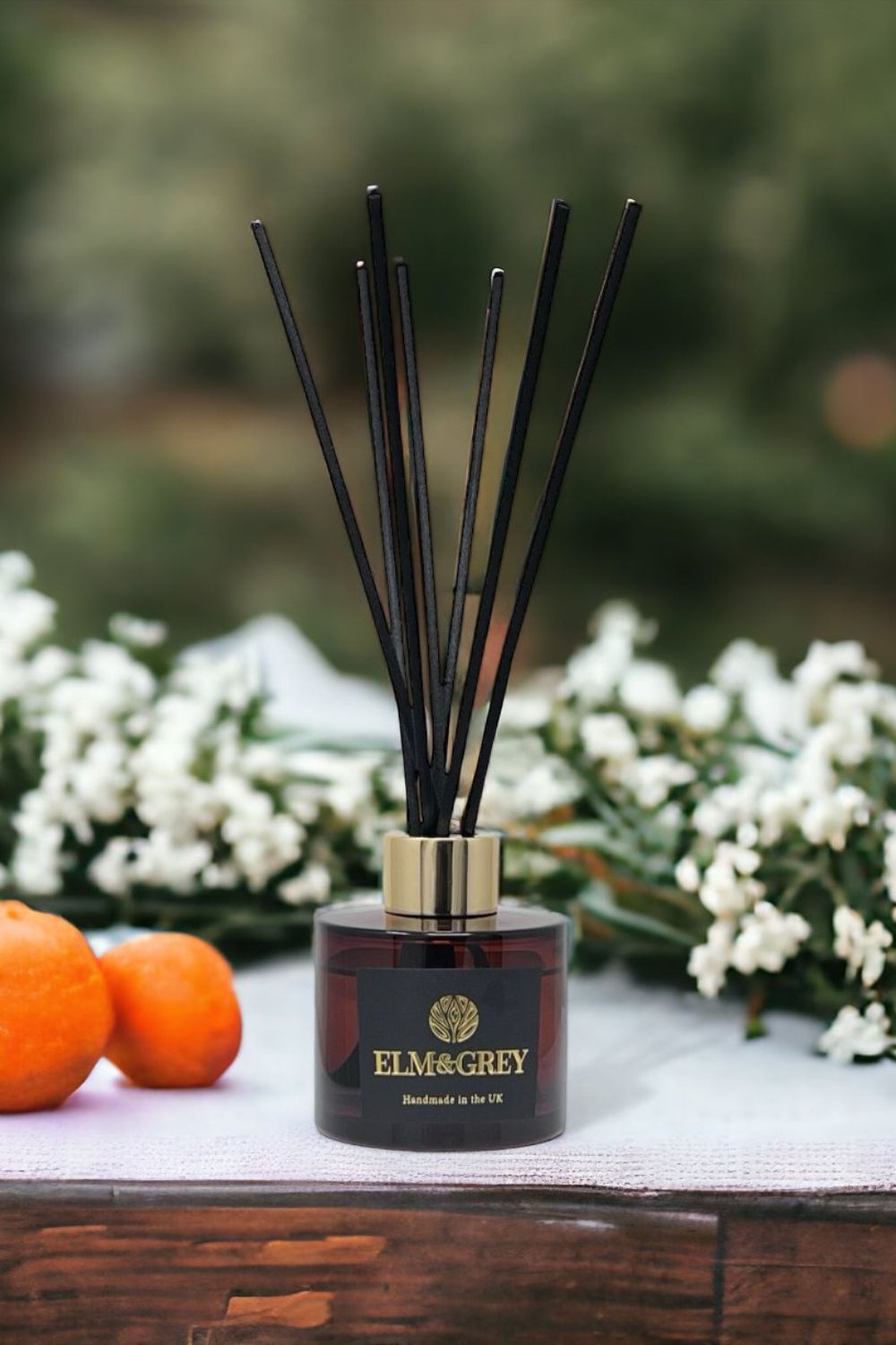 Reed diffuser with black sticks on a wooden surface with oranges and white flowers in the background