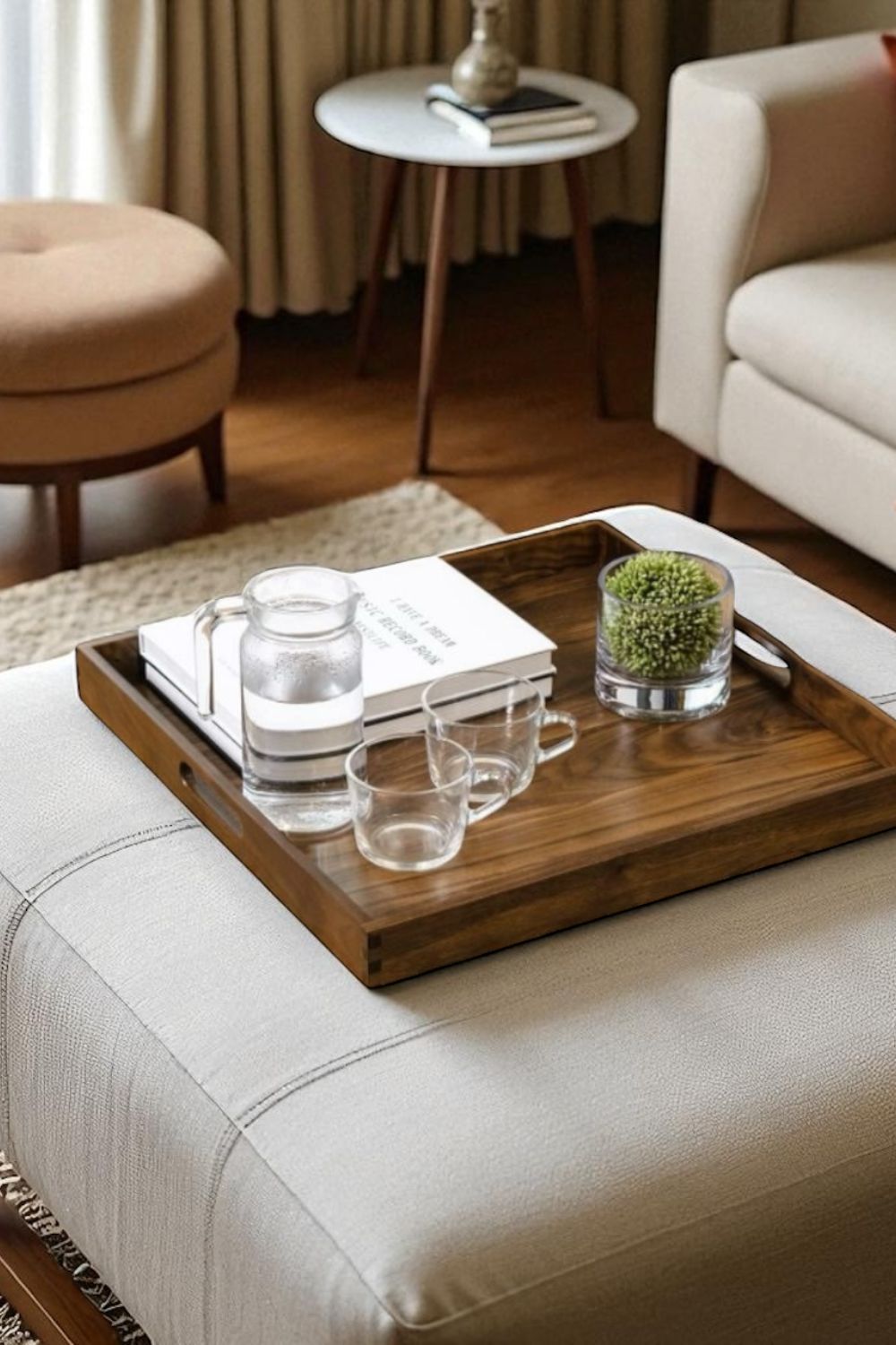 Wooden tray with glasses, water, and a book on a beige ottoman in a living room.