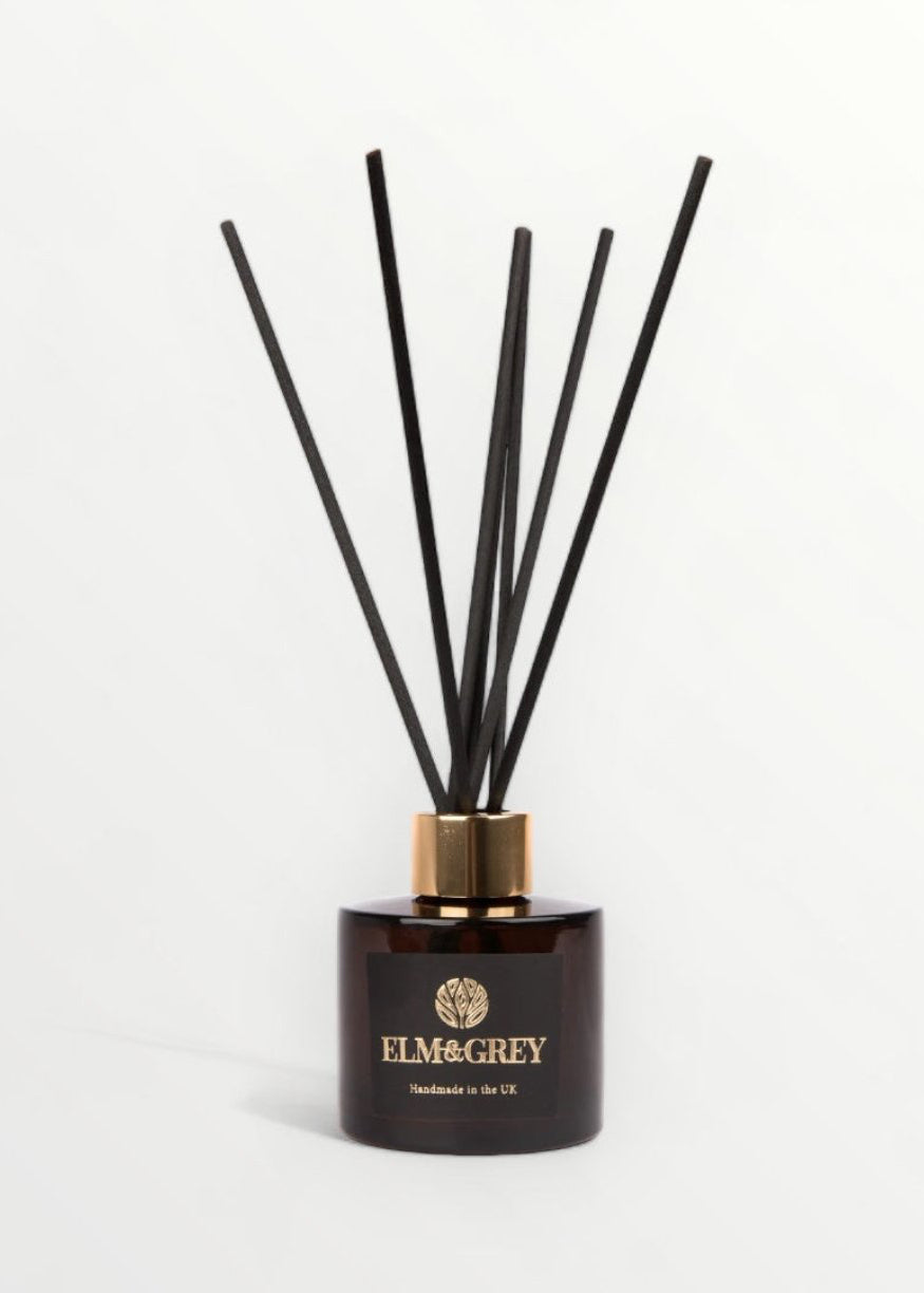 A black reed diffuser with orange blossom and juniper berries on a wooden surface surrounded by greenery and white flowers.