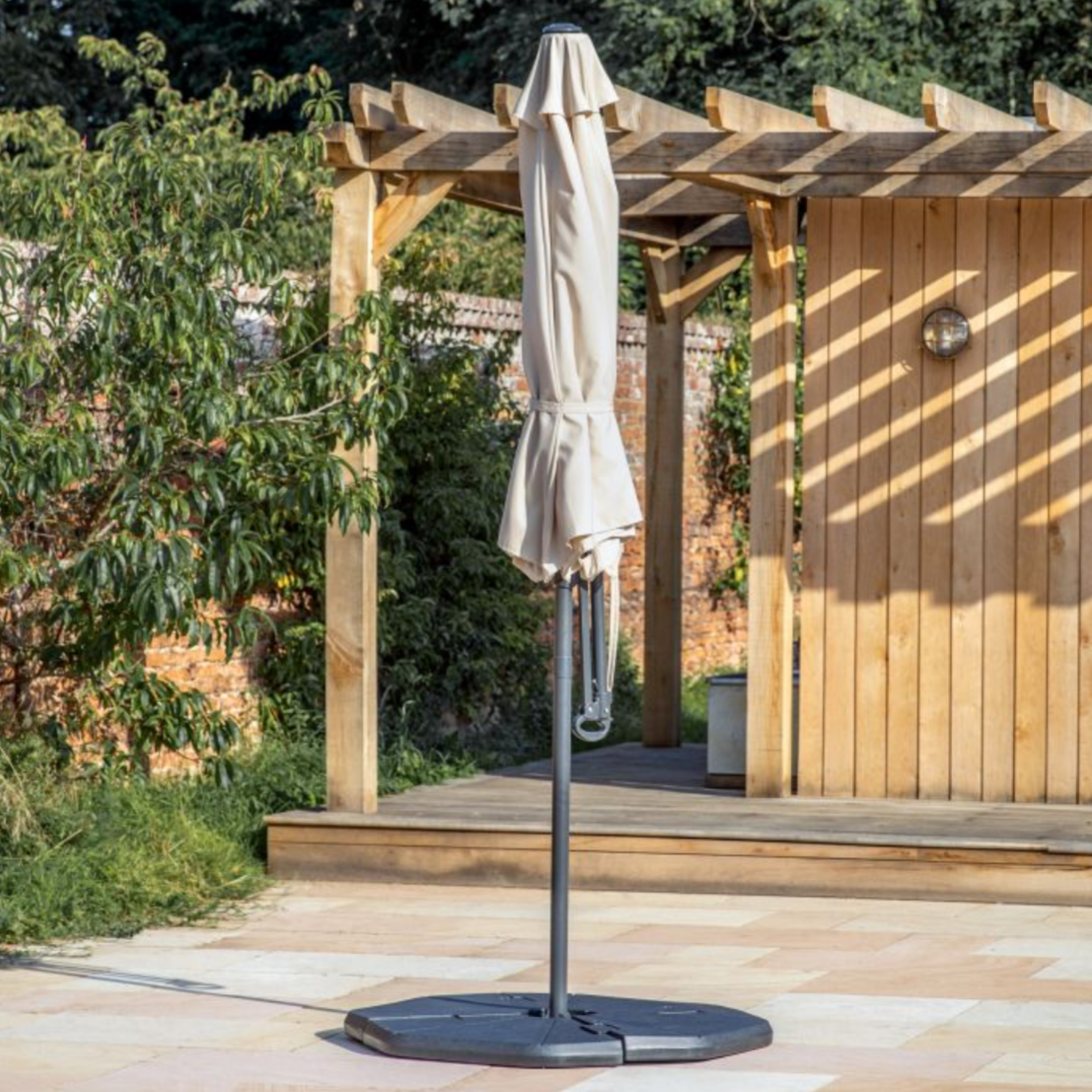 Outdoor umbrella on a stand in front of a wooden pergola with trees and a building in the background.