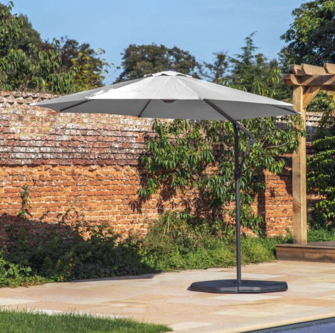 Patio umbrella on a stand in a garden setting with a brick wall and trees in the background.