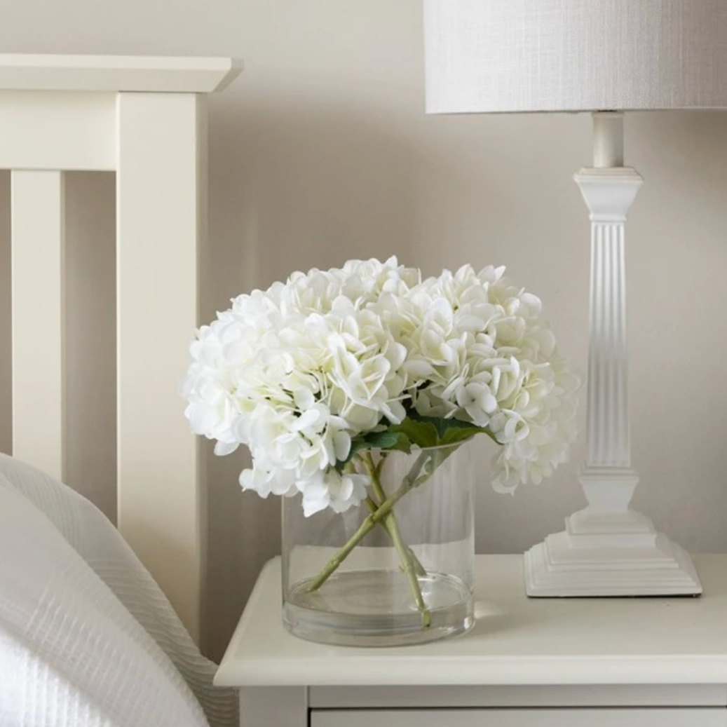 A white hydragea arrangement in a clear cylindrical vase on a side table in a bedroom