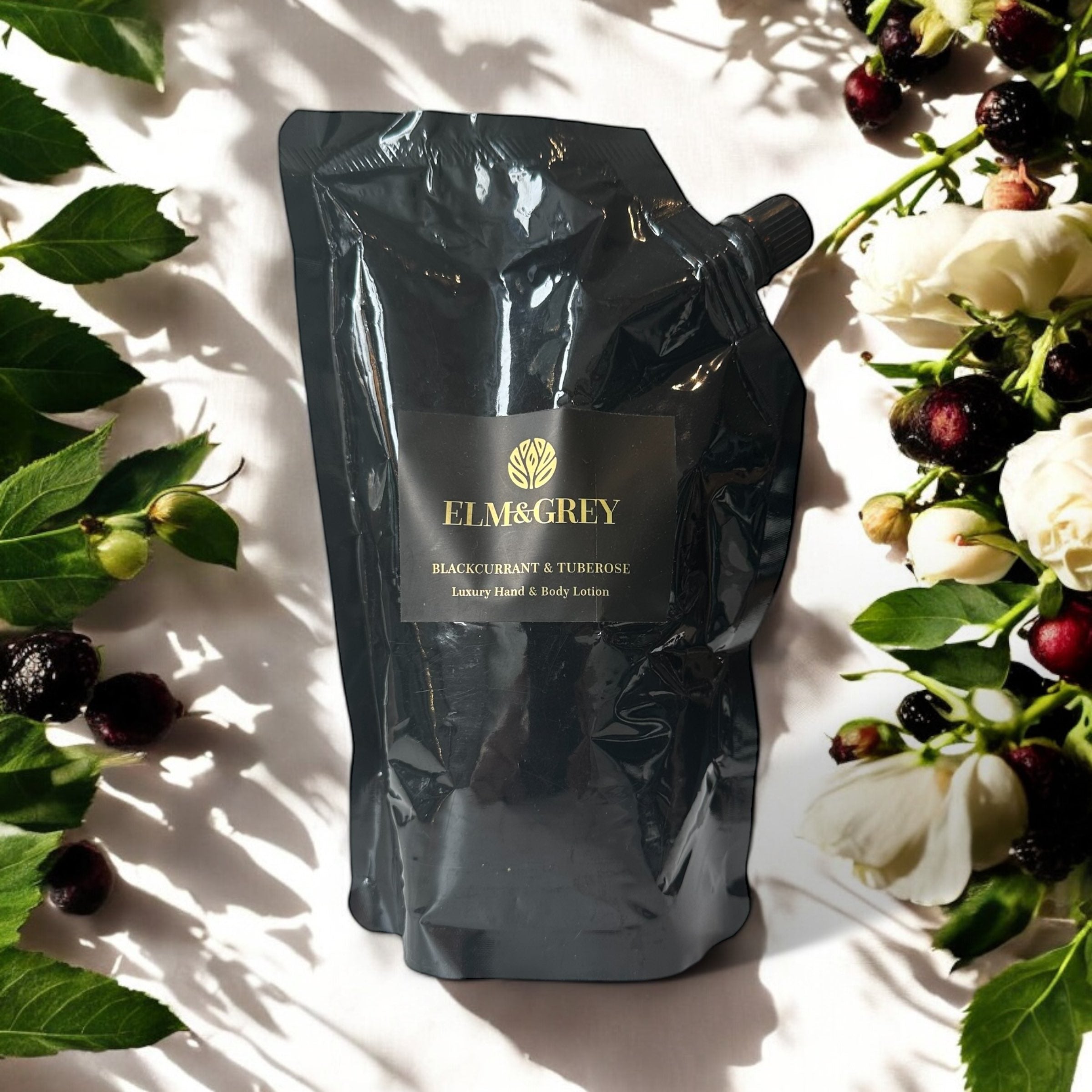 Elm & Grey Blackcurrant & Tuberose Luxury Hand & Body Lotion 450ml Black Refill Pouch sitting on a surface with blackcurrants and roses in the background