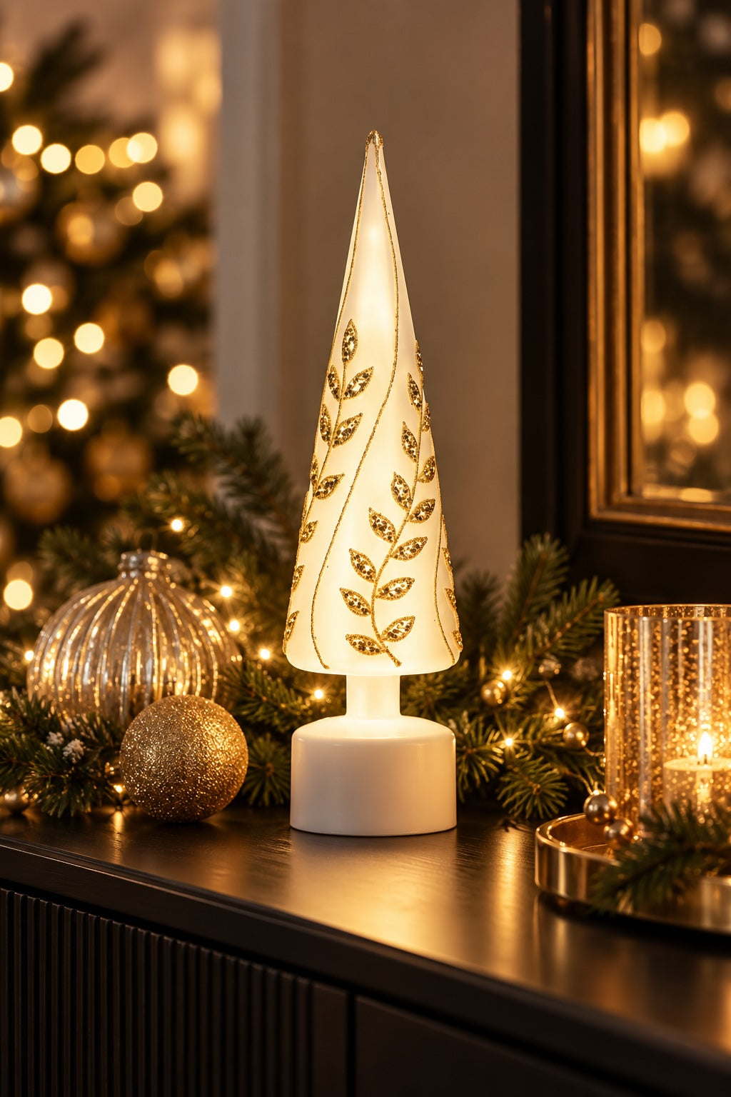 A lit Christmas tree table lamp with a white cylindrical base and a conical shade decorated with gold leaf motifs, styled on a dark mantel with holiday greenery, ornaments, and warm string lights in the background.