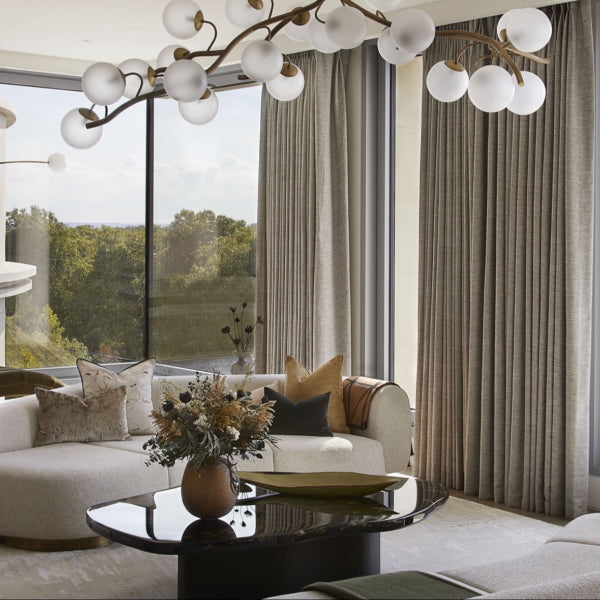 Modern living room with a chandelier, sofa, and large windows with an LSA brown Boulder Vase and dried flowers in the foreground.