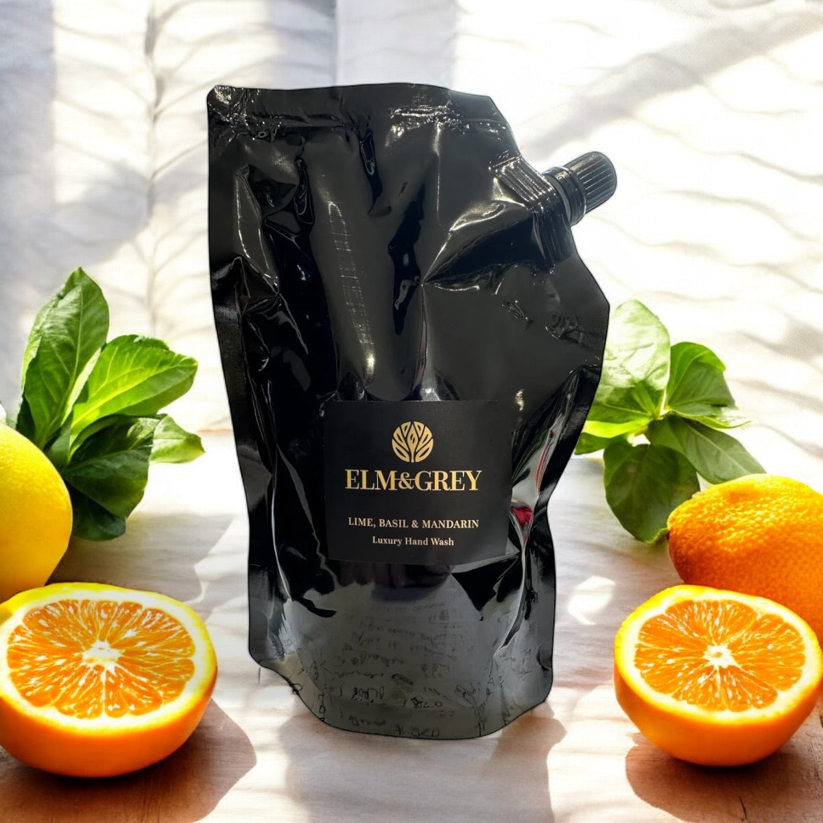 Elm & Grey Dark Lime Basil & Mandarin Luxury Hand Wash 450ml Black Refill Pouch sitting on a surface with limes, basil and oranges  in the background