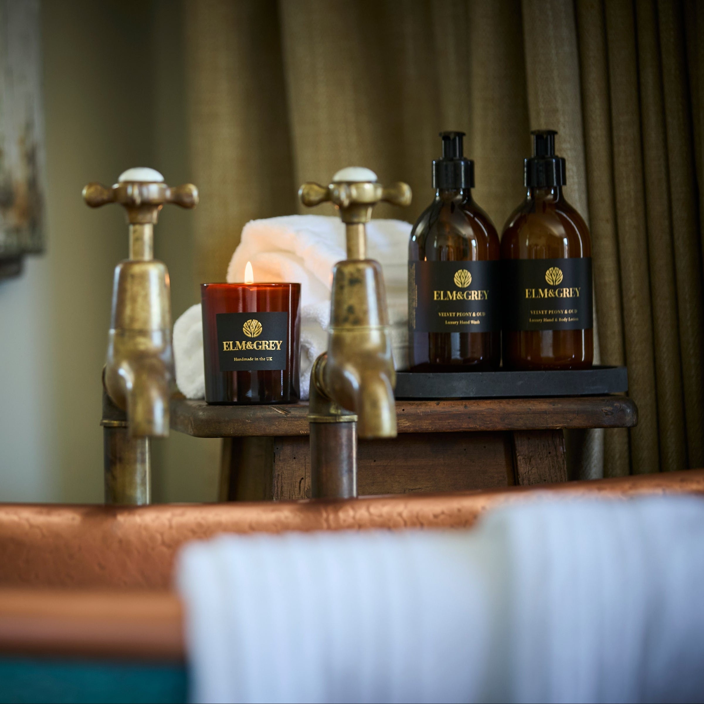 Bathroom setting with Elm & Grey products including bottles, a candle, and brass faucets.