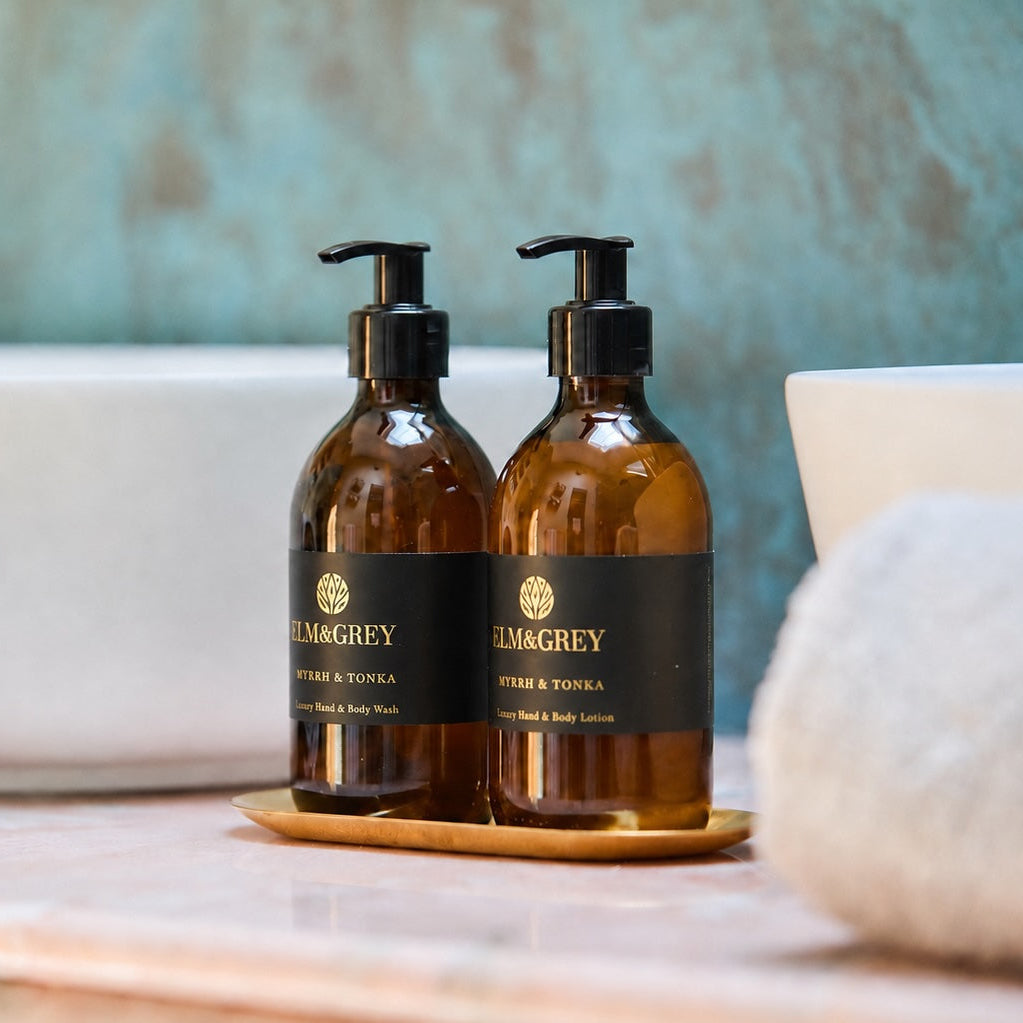 Two Elm & Grey Luxury Hand Wash & Lotion Amber Glass bottles with pumps, set on a marble countertop with rolled towels and a washbasin in the background.