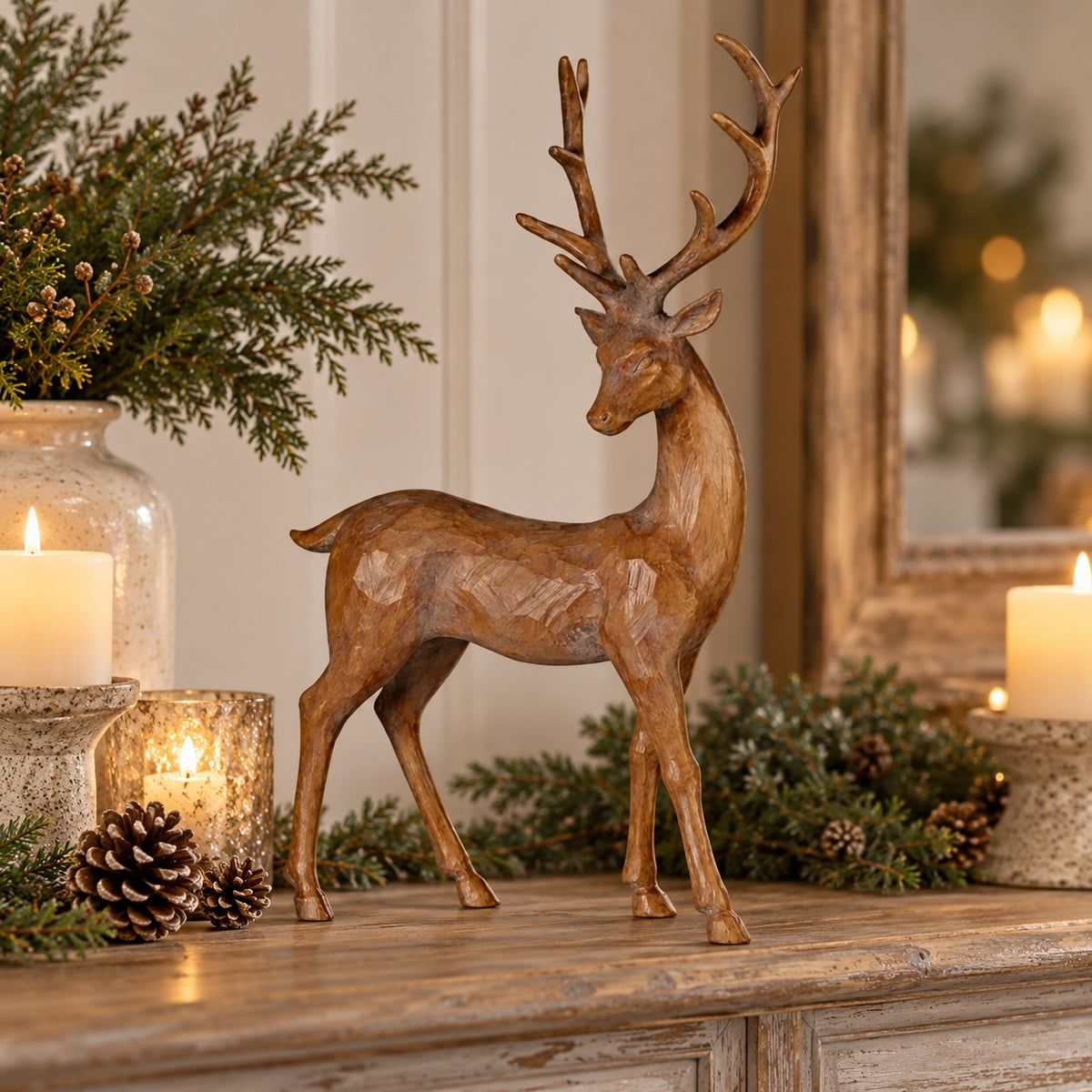 A decorative wooden stag figurine with antlers stands on a rustic wooden console table, surrounded by lit candles, pinecones, evergreen branches, and a mirror in a warm holiday interior setting.