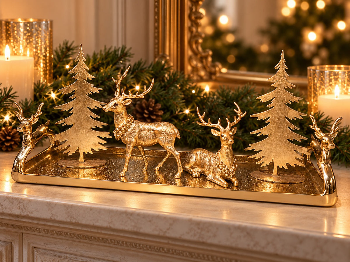 A gold-toned Christmas decorative tray set on a marble mantel, featuring two deer figures, two small tree silhouettes, and lit candles in the background with a blurred festive tree and mirror.