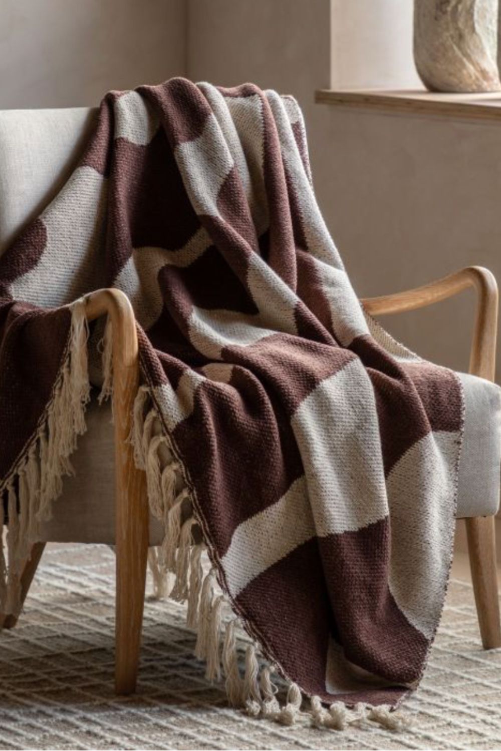 Brown and cream abstract striped blanket with tassels draped over a wooden chair in a cosy room.
