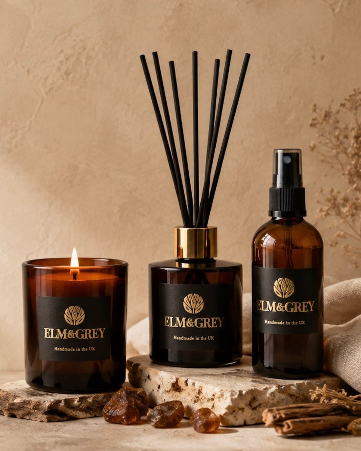 A home fragrance set by ELM&GREY, including an amber glass candle, a reed diffuser with black reeds, and an amber glass spray bottle, styled on a stone slab with dried botanicals against a textured beige background.