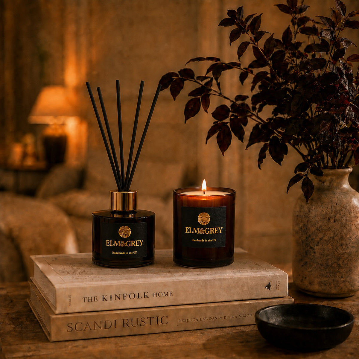 Elm & Grey amber reed diffuser and lit candle styled in a warm, cosy living room with soft ambient lighting