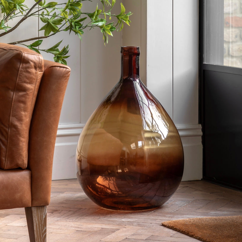Large amber glass vase on a wooden floor next to a brown leather armchair.