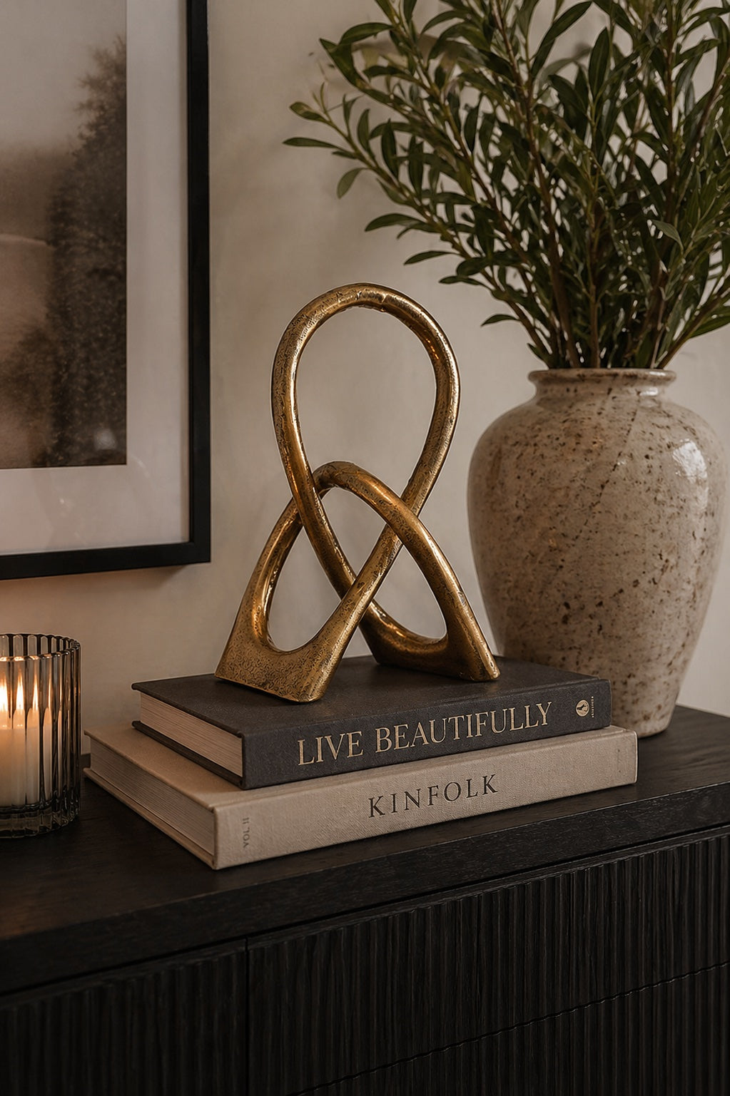 Antique gold knotted loop decorative sculpture on stacked Live Beautifully and Kinfolk books on a dark fluted sideboard with lit tealight and olive branches — Elm & Grey