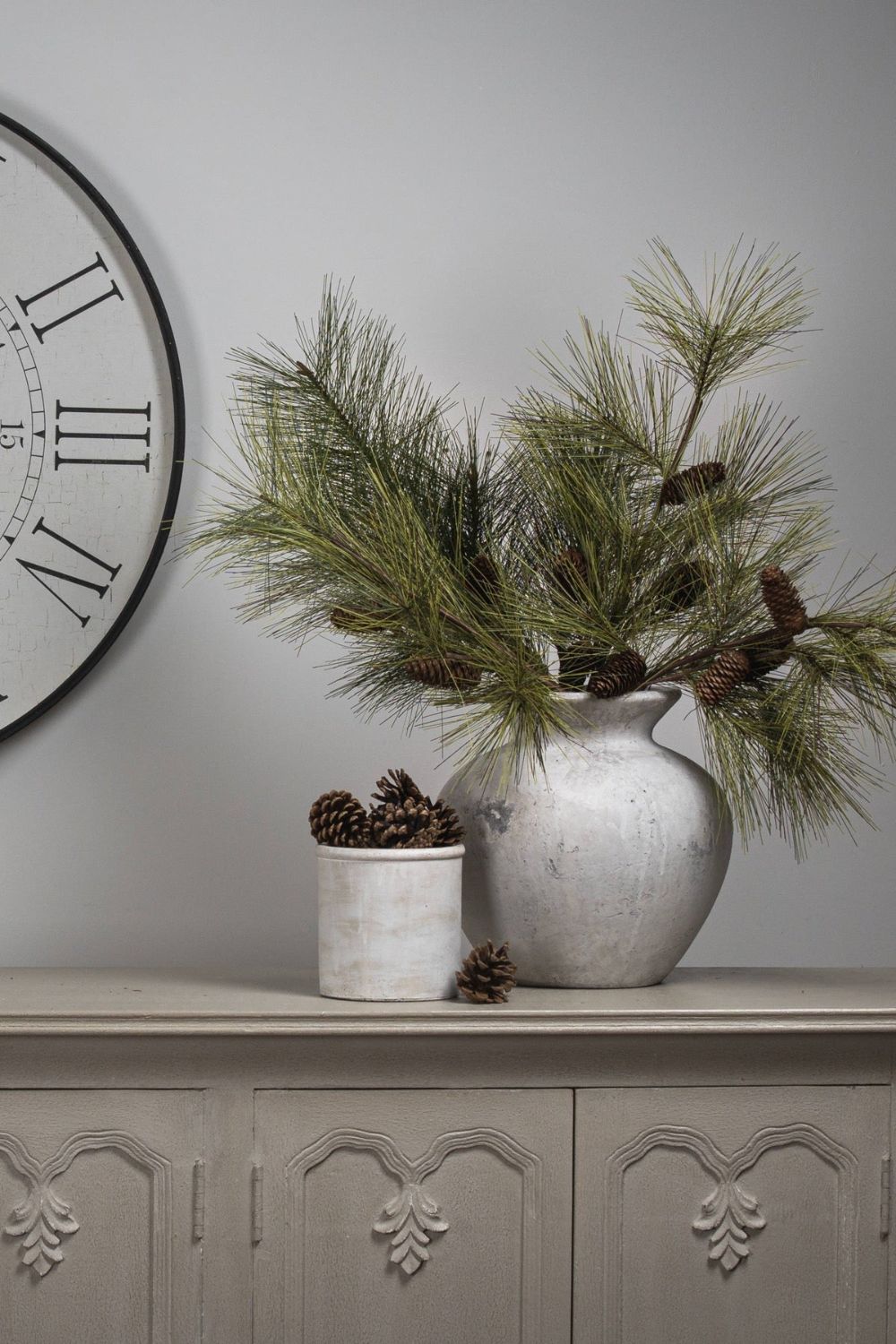Decorative setting with a vase of greenery, small pot, and clock on a gray surface.