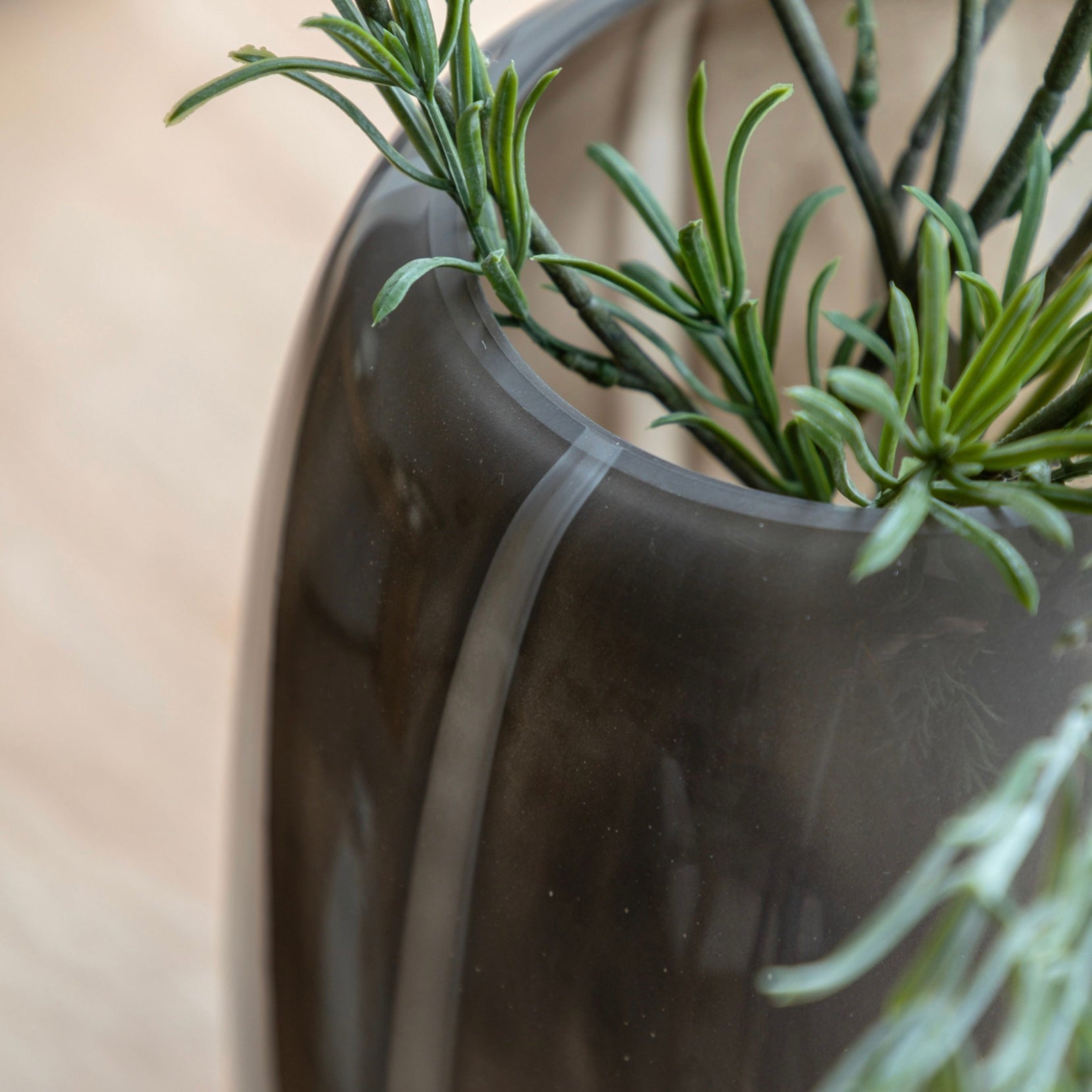 Close-up of a taupe glass vase with green plants against a neutral background