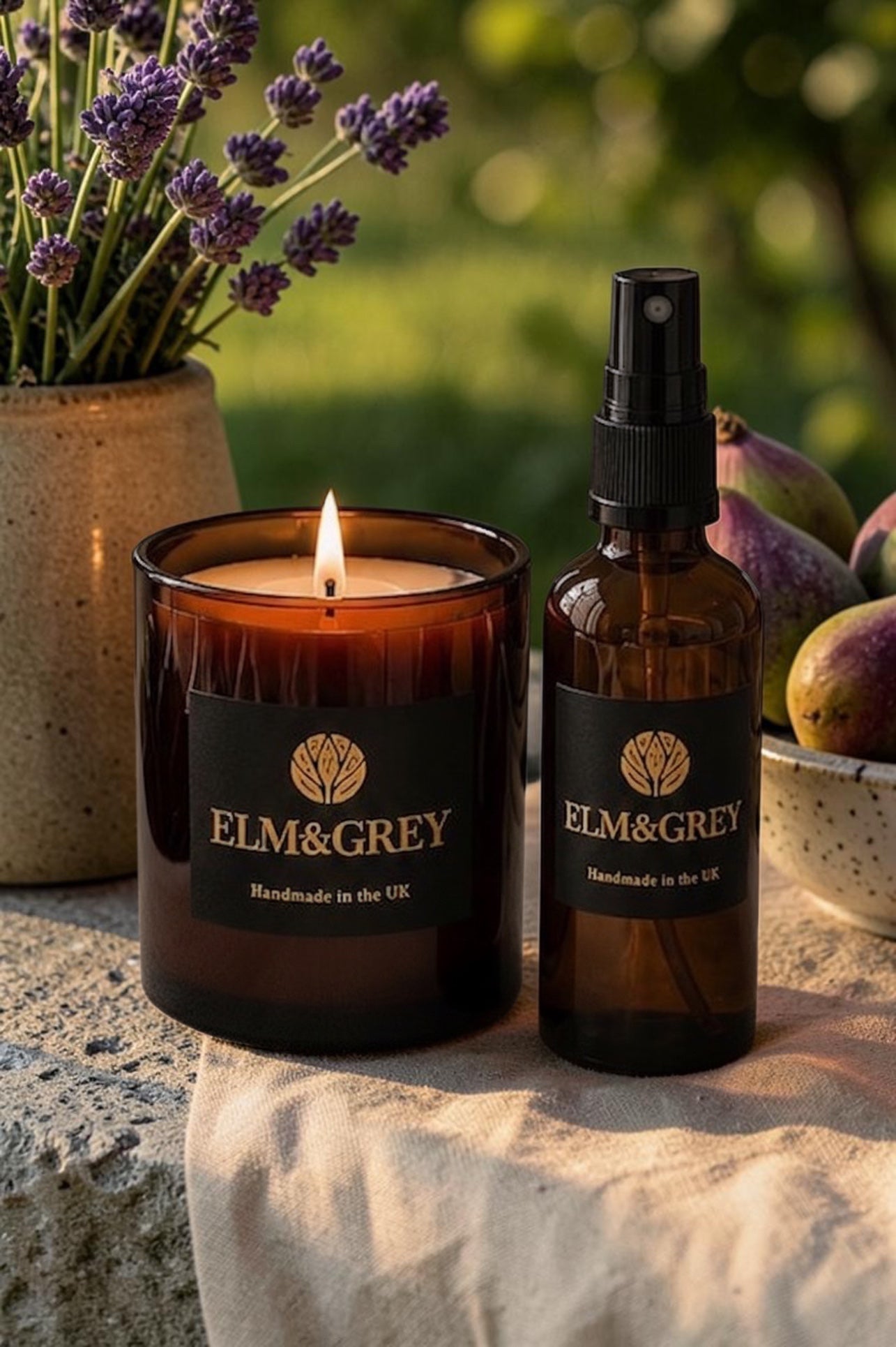 A lit amber glass jar candle and a matching amber spray bottle with black labels reading ELM&GREY, styled outdoors on a stone surface with lavender and figs in the background.