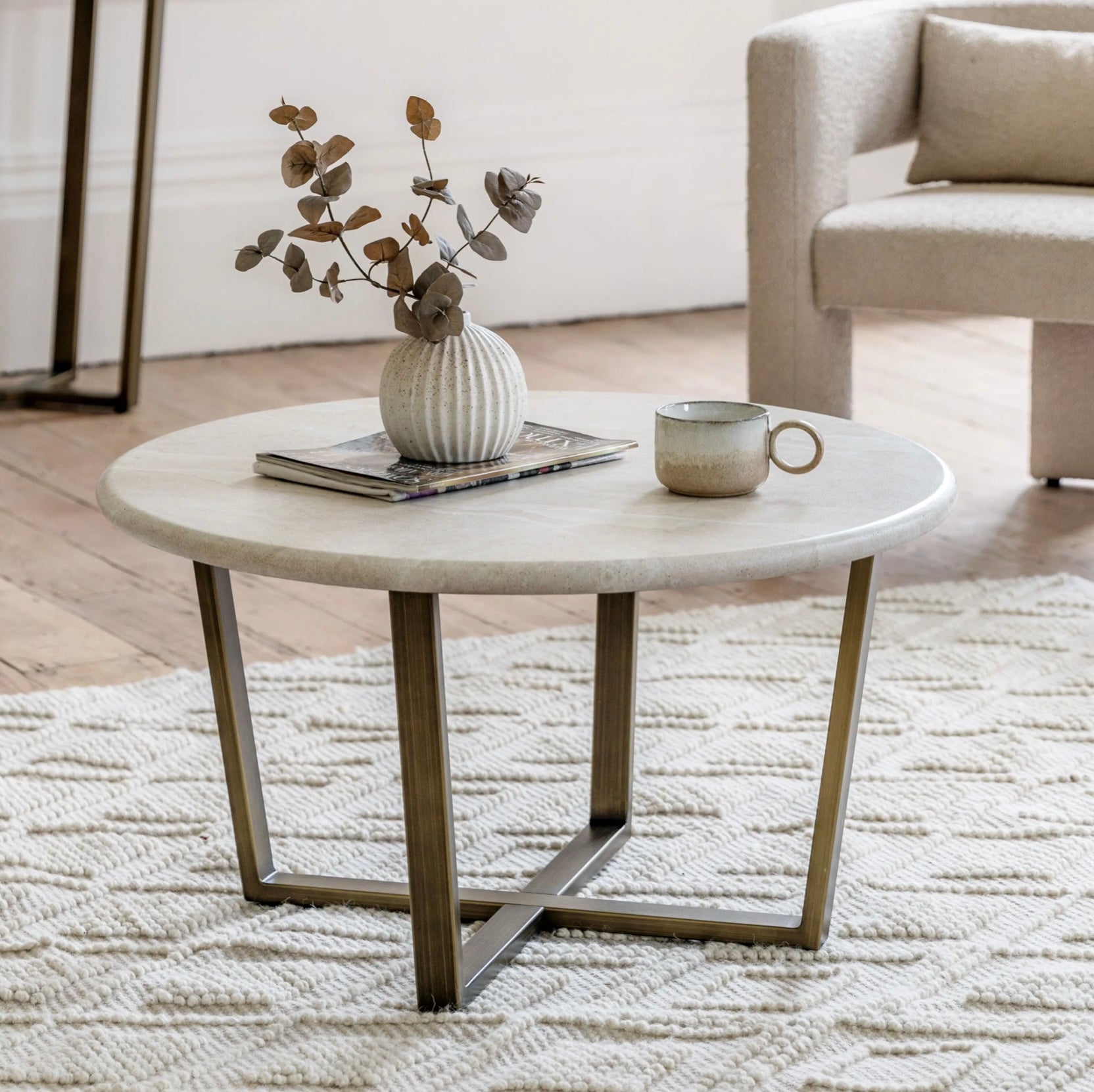 Travertine effect round coffee table with brass base on a rug with armchair