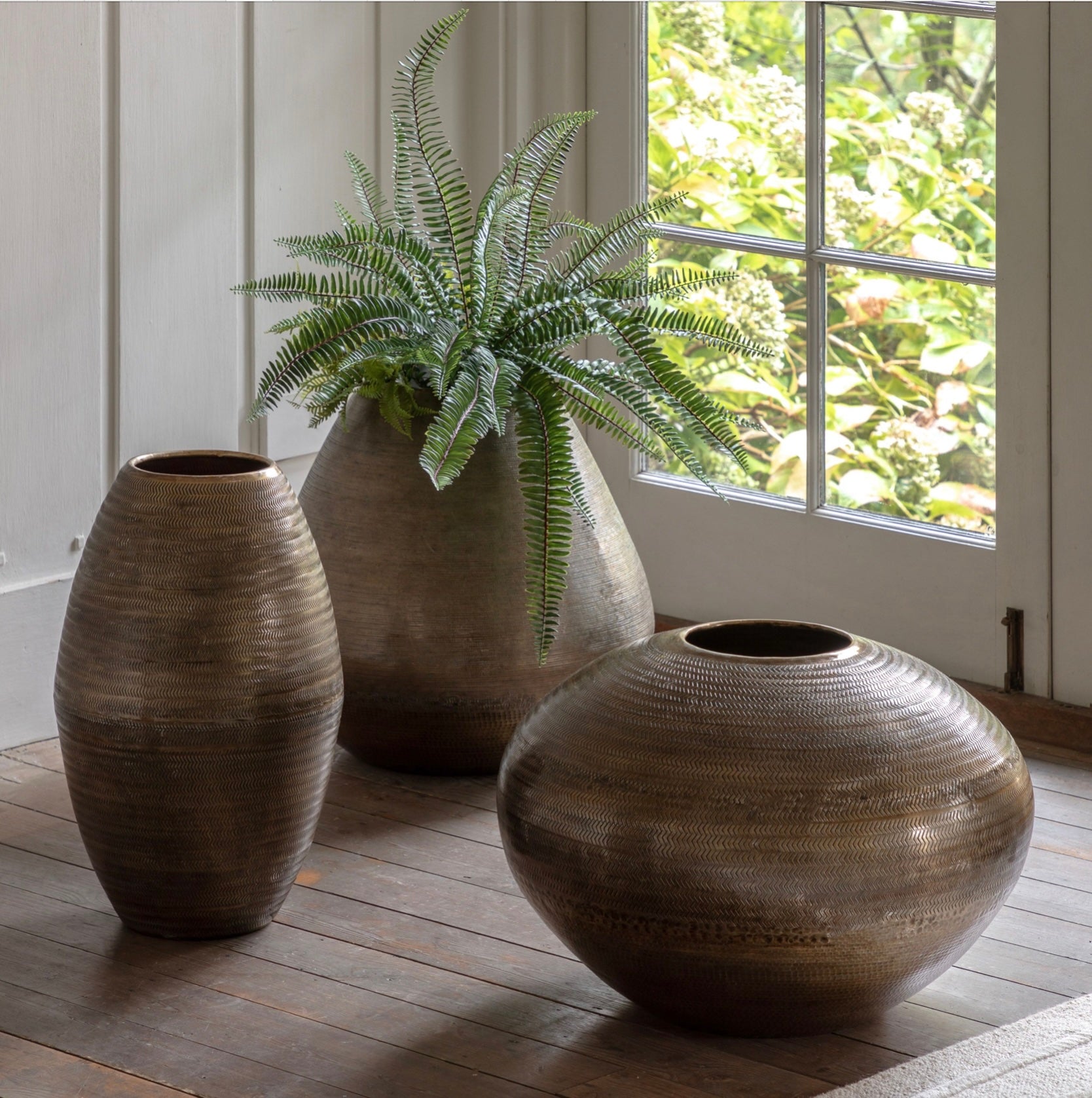 Three decorative antiqued brass vases on a wooden floor with a plant in the background.