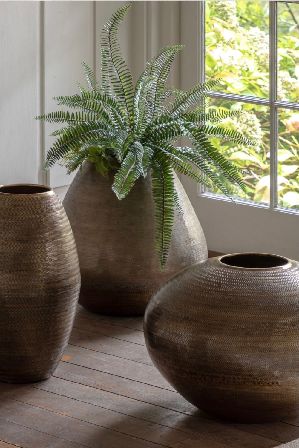 Fern in a textured pot on a wooden floor with large decorative antiqued brass vases.