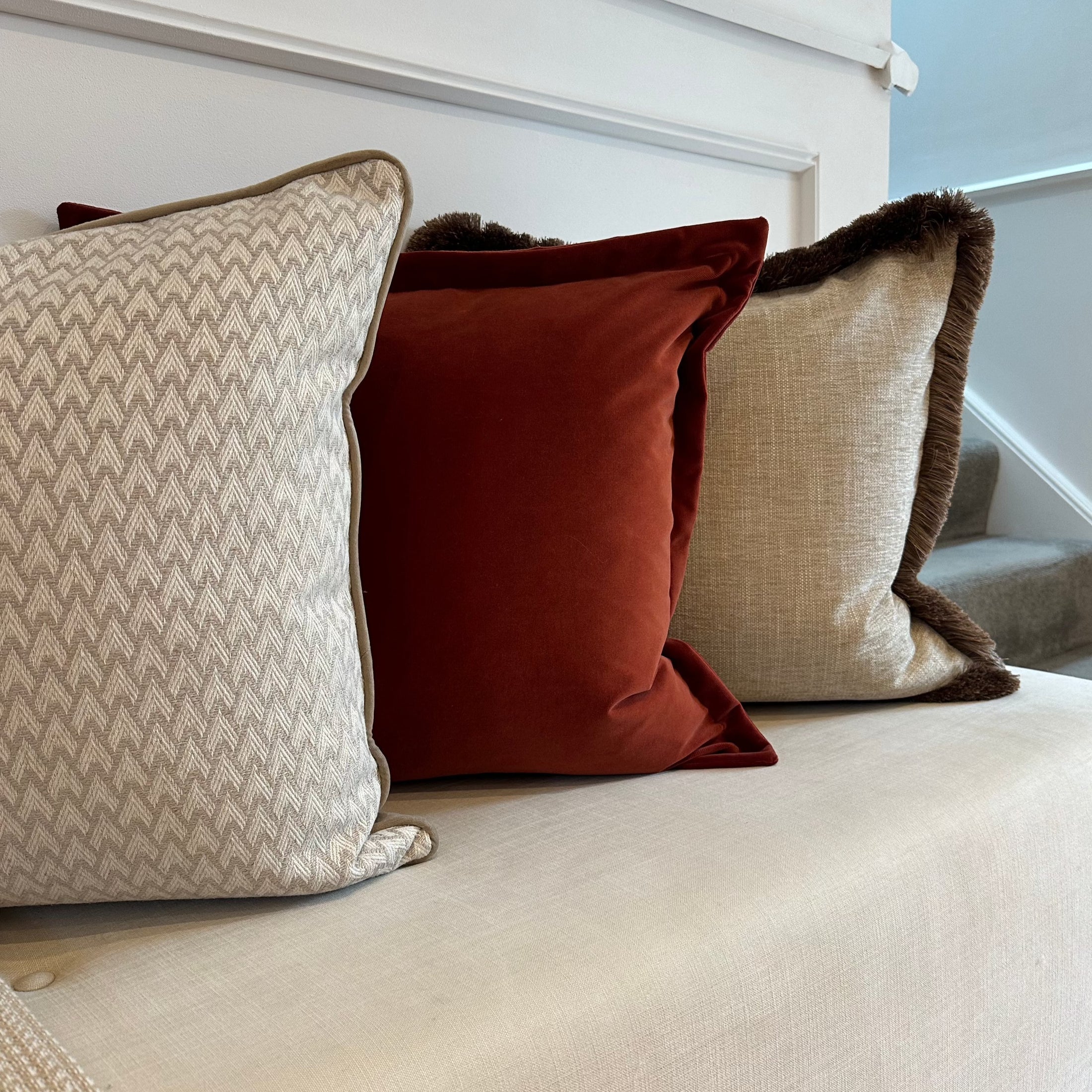 Three cushions in shades of beige, orange, and gray, with different textures and trim, arranged on a sofa.