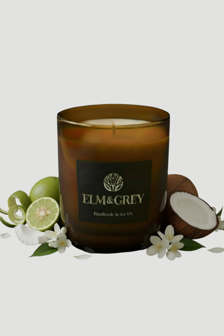Amber glass candle with 'Elm & Grey' branding surrounded by limes, coconuts, and flowers on a light grey background
