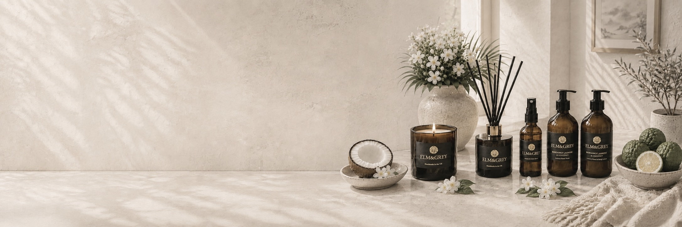 Bergamot, Jasmine & Coconut Home Fragrance products by Elm & Grey  including amber glass bottles, pump dispensers, and a reed diffuser, styled on a textured beige surface with a candle, flowers, and citrus slices under soft window shadows.