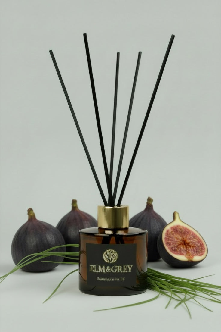 Reed diffuser with black sticks and a gold cap on a grey background with figs and green leaves.