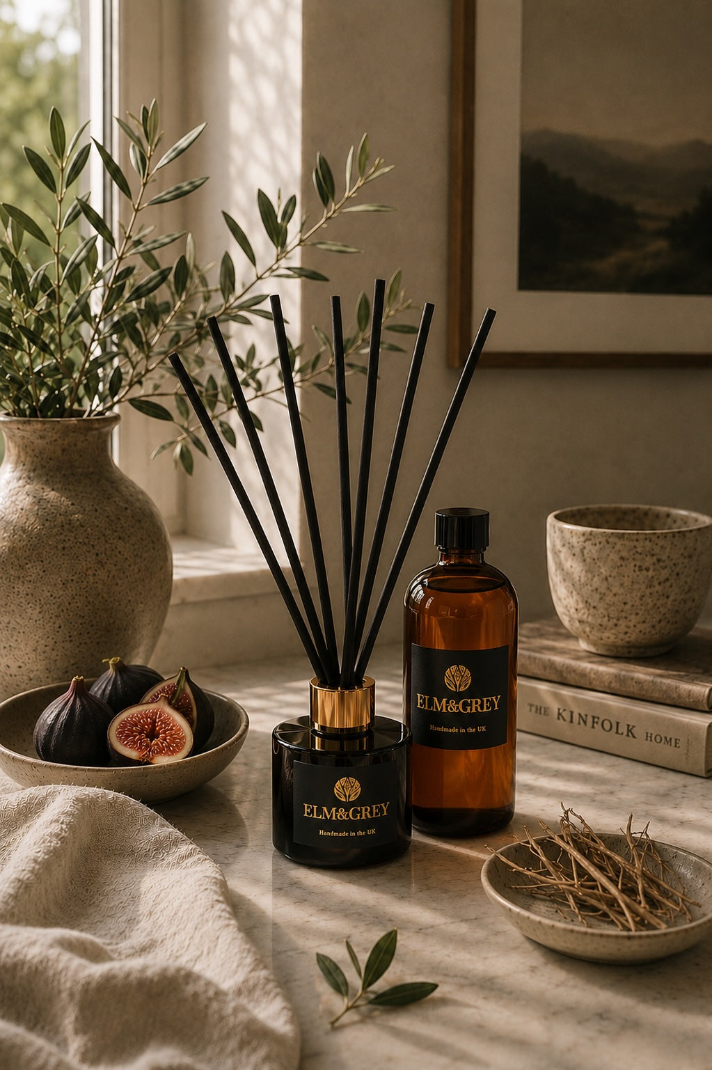 Elm & Grey Black Fig & Vetiver reed diffuser with diffuser refill bottle styled on a marble countertop with fresh figs, olive branches and natural ceramics. Luxury amber glass home fragrance handcrafted in the UK, creating a warm and sophisticated atmosphere in the home.
