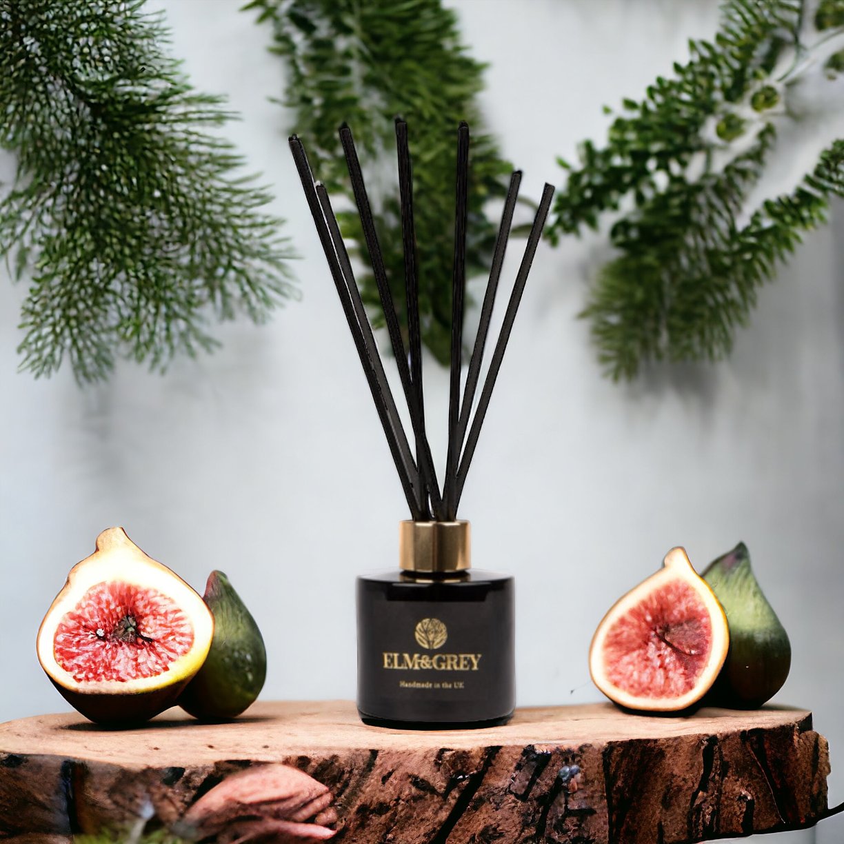 A reed diffuser with black sticks in a matt black glass bottle, flanked by two halves of a fig, on a wooden surface with greenery in the background.