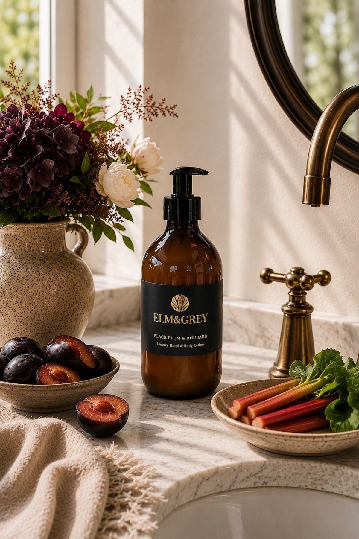Elm & Grey Black Plum and Rhubarb luxury hand and body lotion on marble bathroom surface with black plums, rhubarb stalks and dark floral arrangement beside brass tap