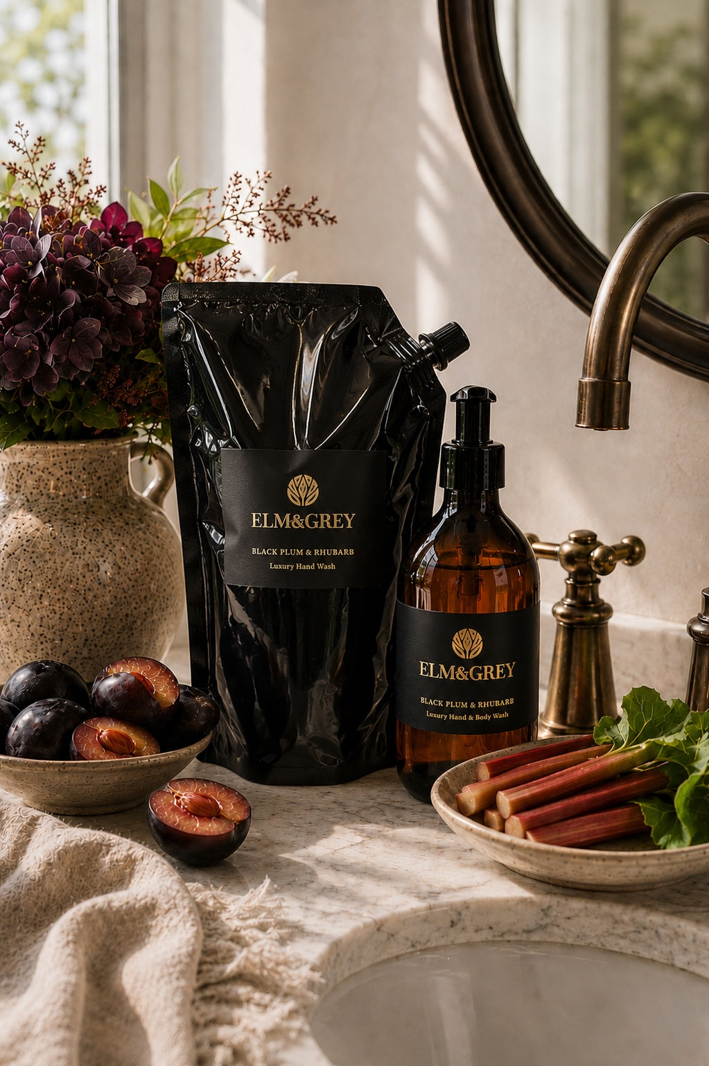 Elm&Grey black plum and rhubarb hand wasn't refill pouch and bottle on a bathroom counter with fruits and flowers.