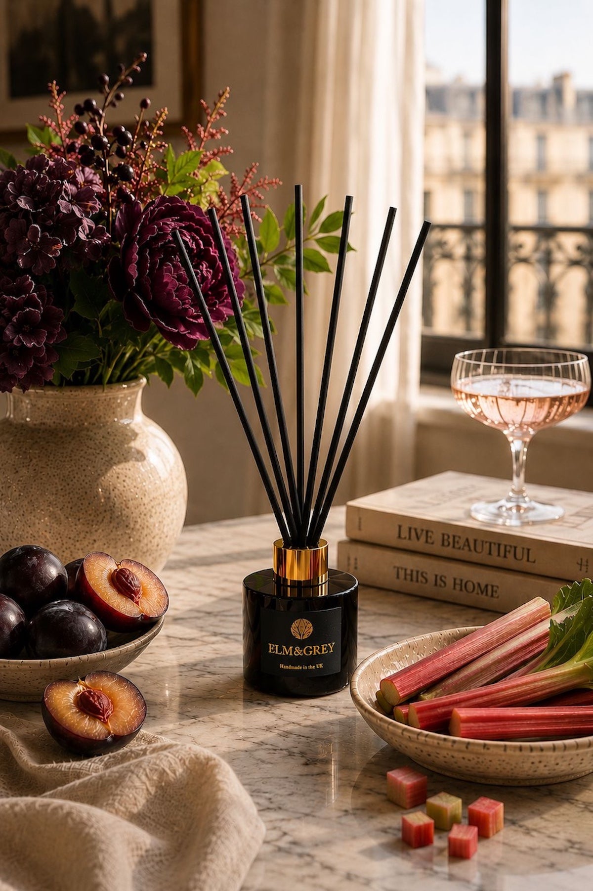 Elm & Grey Black Plum and Rhubarb reed diffuser on marble surface with deep purple peonies, black plums, rhubarb and rosé glass in a Parisian window setting