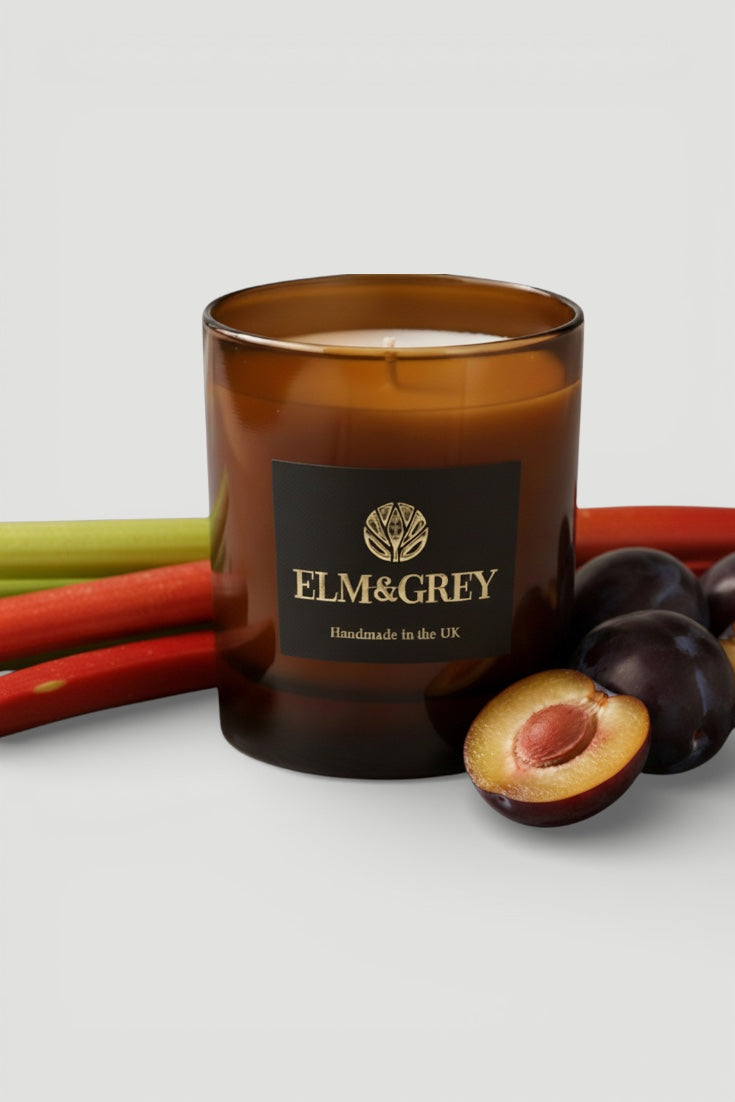 Candle with Elm & Grey branding surrounded by plums and a green stick on a light background