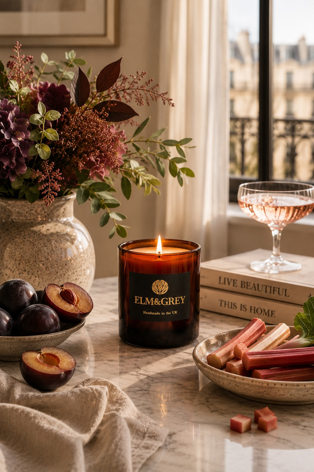 Black Plum and Rhubarb scented candle by elm and grey in a softly lit lounge setting styled with flowers and fruit on a marble surface.