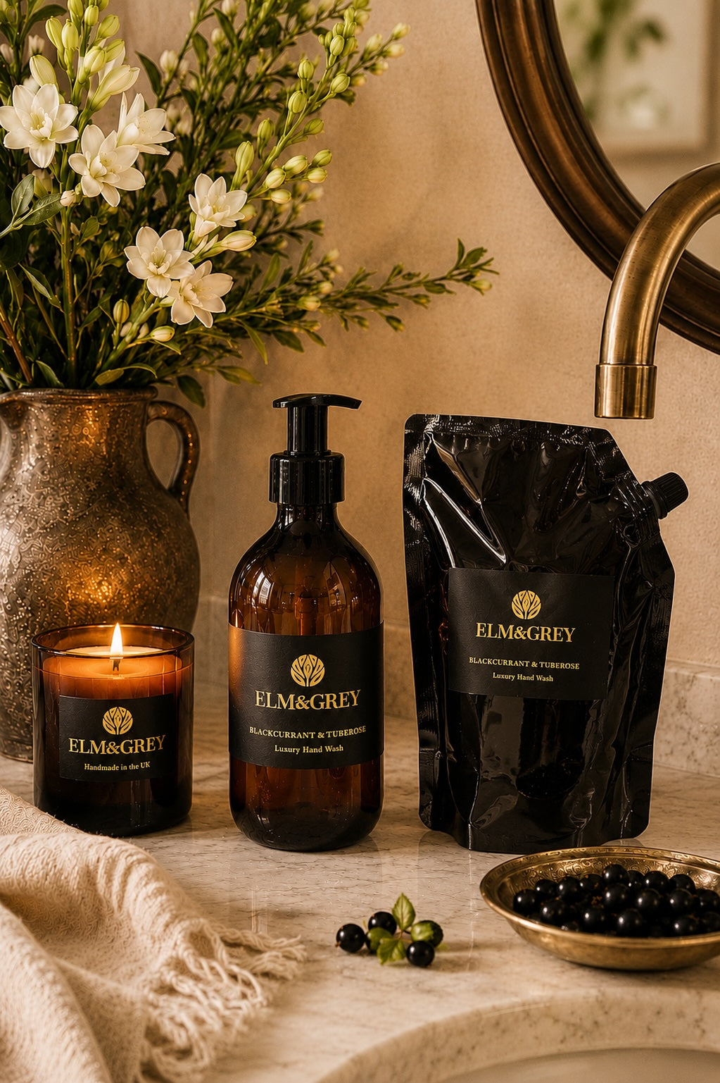 Blackcurrant and tuberose hand and body wash refill pouch, bottle and scented candle by Elm and Grey. Styled on a marble sink countertop with florals and fruit.