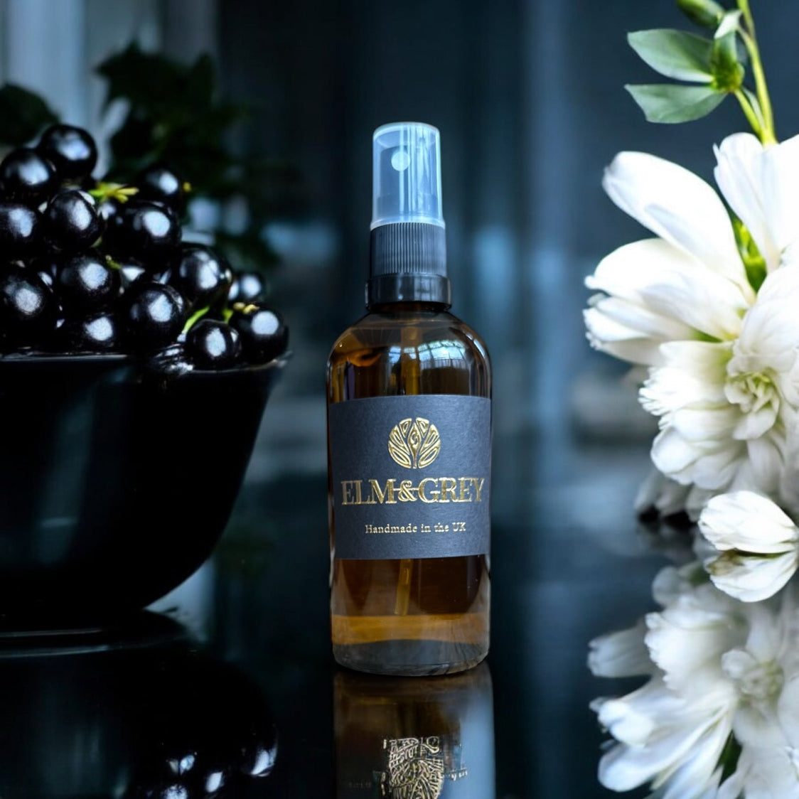 A bottle of Blackcurrant and Tuberose Room Spray with a spray nozzle, placed on a surface reflective with a floral arrangement and fruits in the background.
