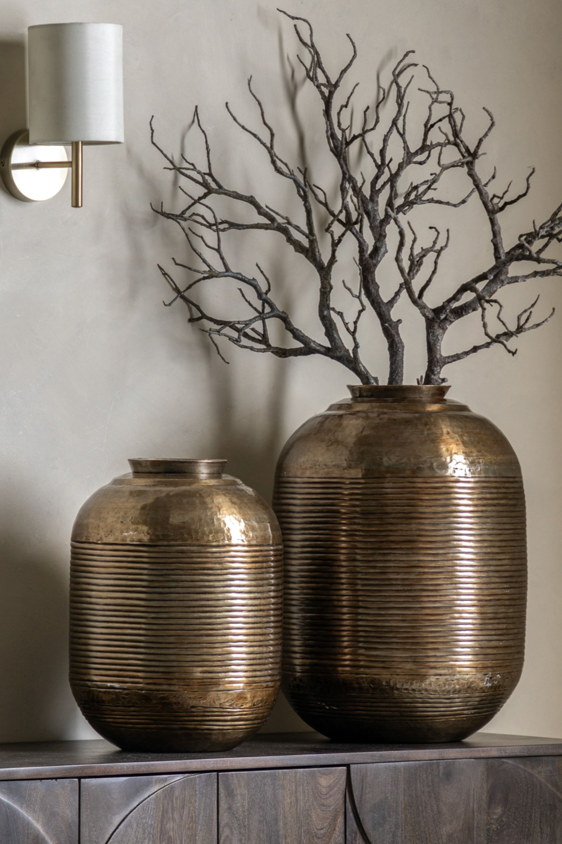 Two bronze vases with textured surfaces on a wooden surface against a neutral wall.