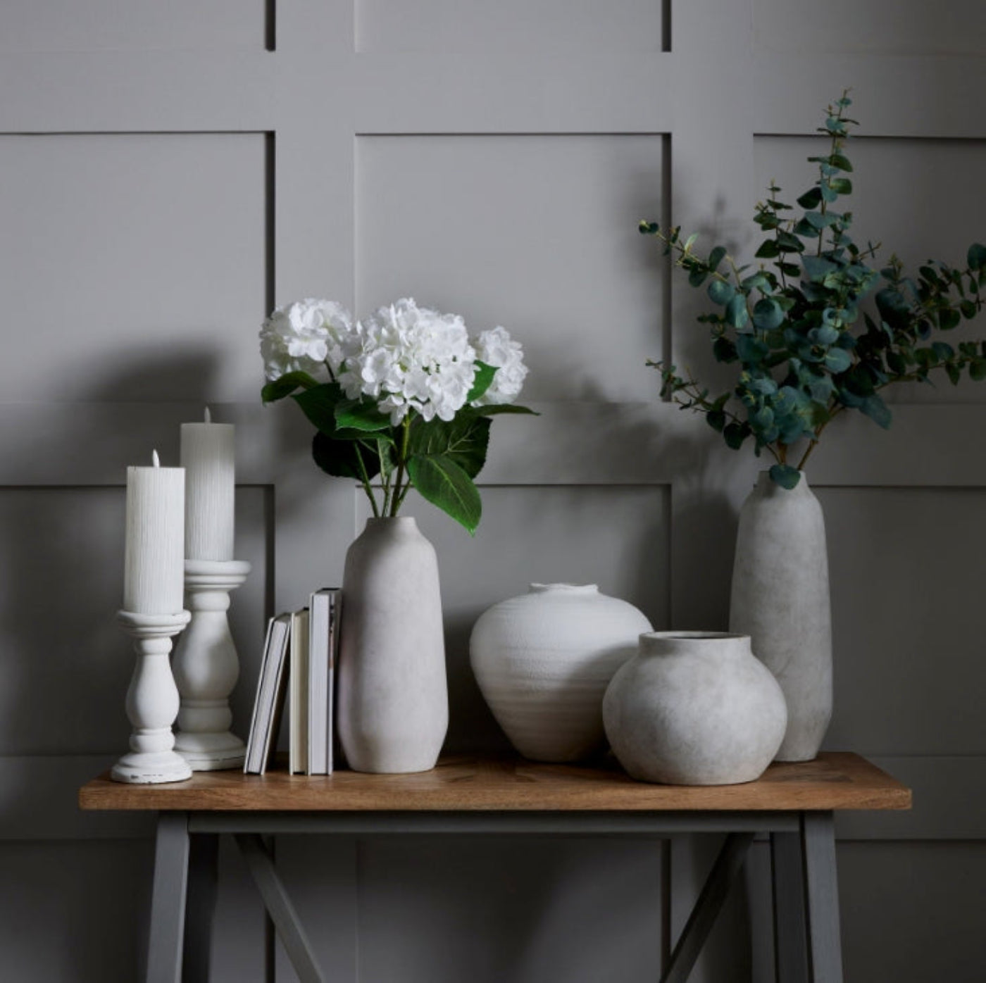 Decorative setting with vases, candles, and books on a wooden table against a grey paneled wall.