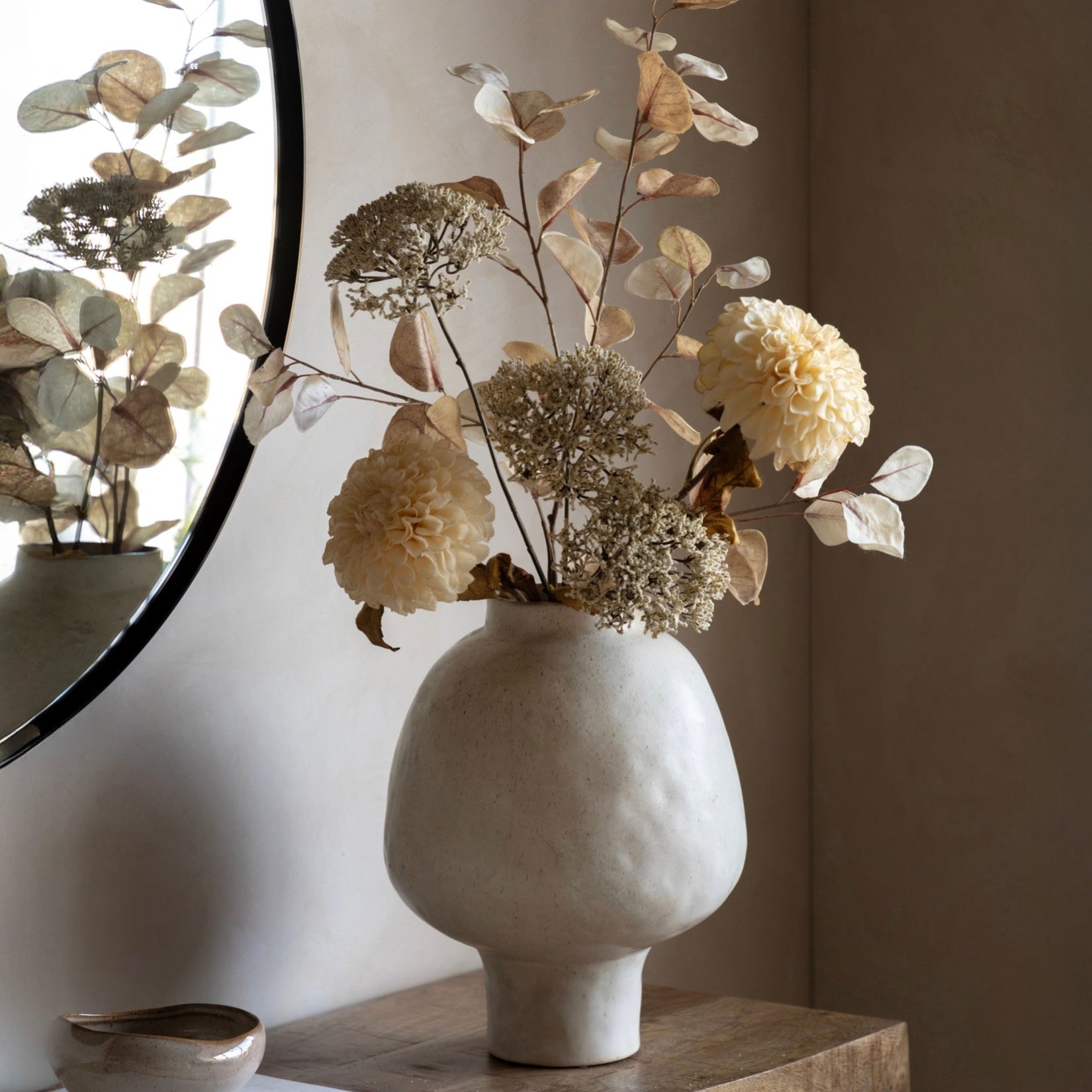 White textured vase with dried flowers on a wooden surface, mirror reflecting floral arrangement.