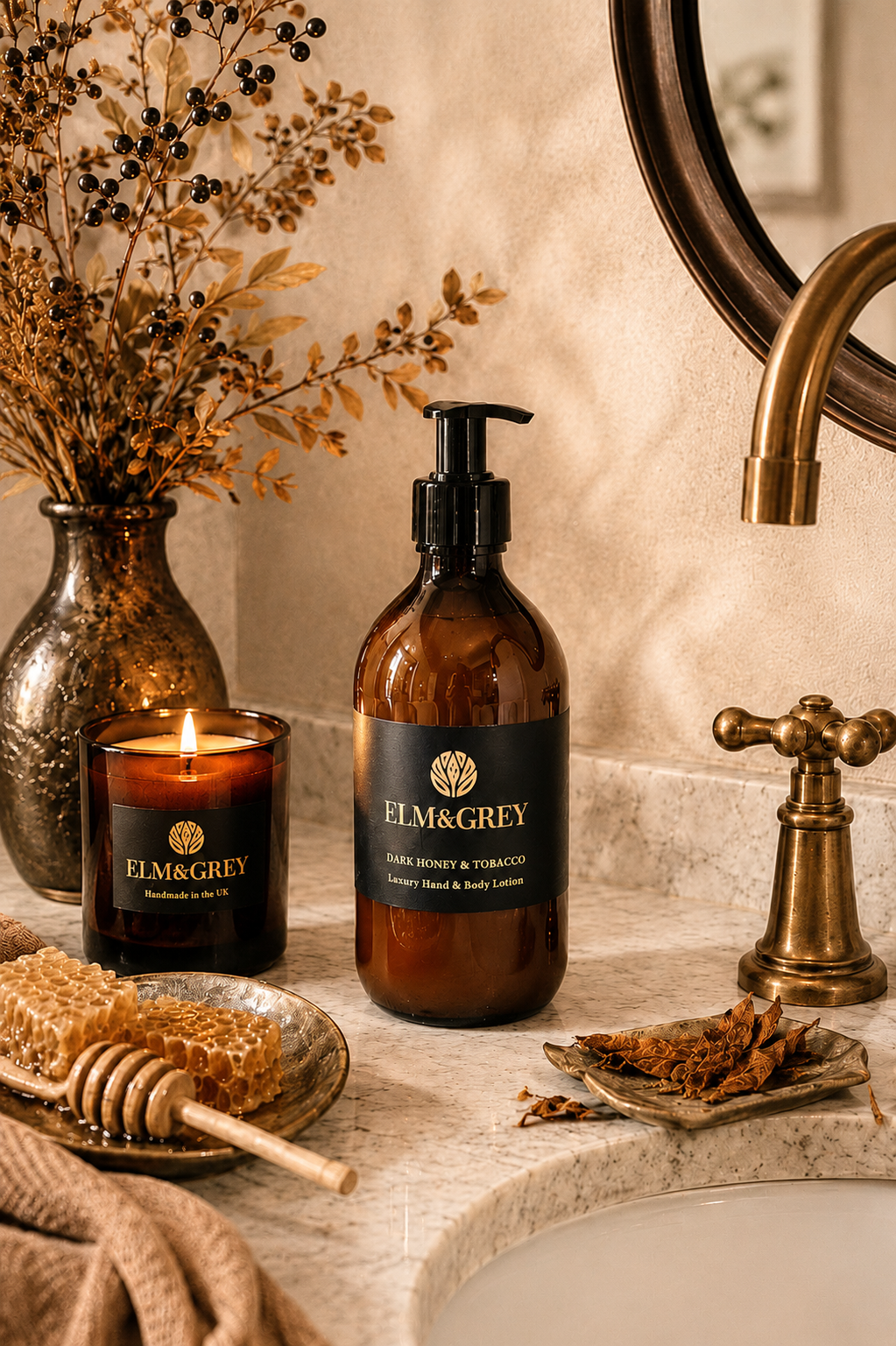 Elm & Grey Dark Honey and Tobacco luxury hand and body lotion on marble bathroom surface with lit candle, honeycomb, dark berry branches and tobacco pieces beside brass tap