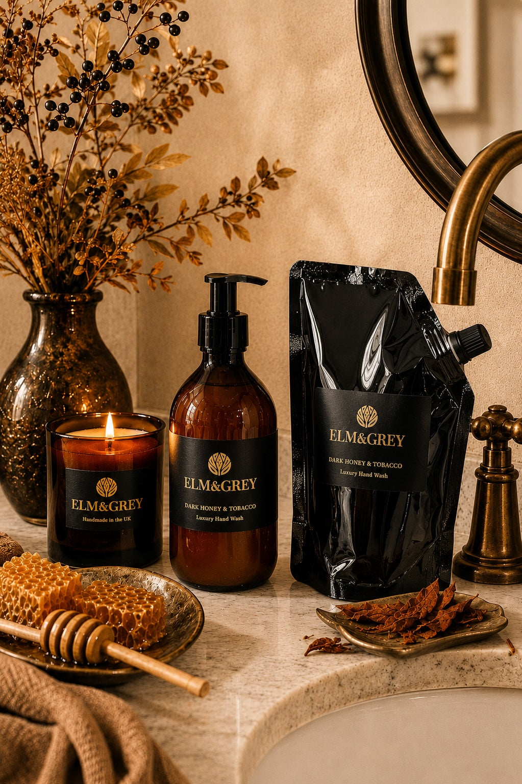 Dark honey and tobacco fragranced candle, hand and body wash and refill pouch by Elm and Grey. Styled on a marble vanity unit with botanicals.