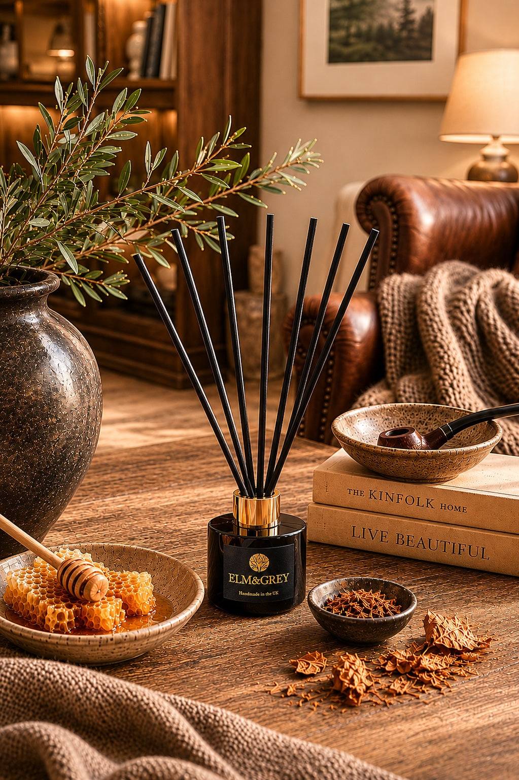 Elm & Grey Dark Honey and Tobacco reed diffuser on a dark wooden surface styled with honeycomb, tobacco and clove pieces, olive branches and Kinfolk books in warmly lit living room.