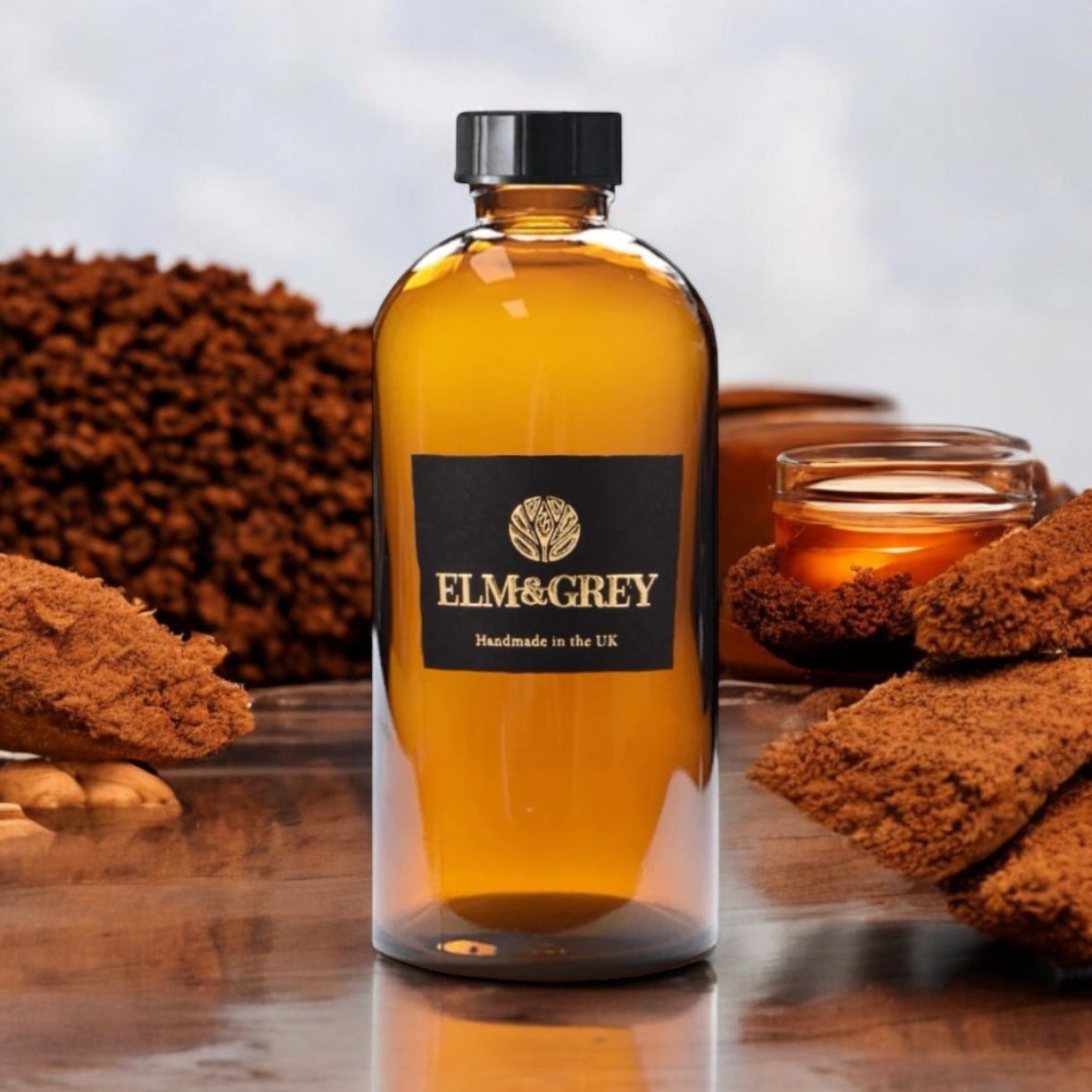 A clear glass bottle of Dark Honey and Tobacco diffuser refill with a yellow-hued liquid, branded Elm & Grey, accompanied by what appears to be tobacco leaves and a honeycomb.