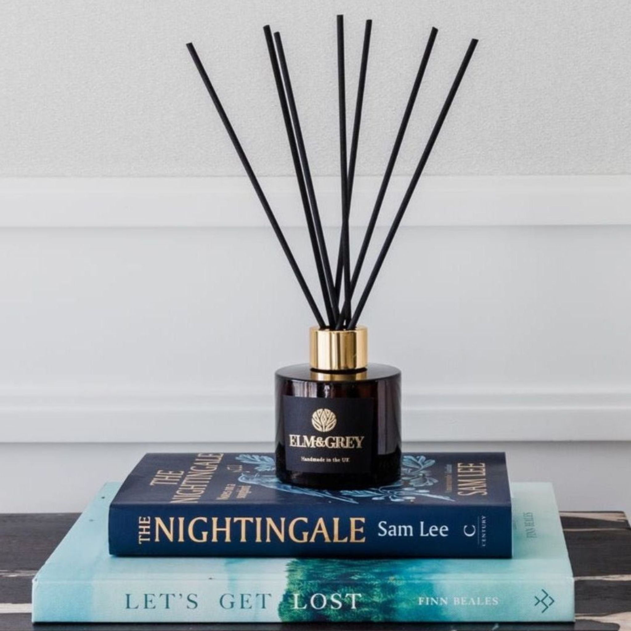Reed diffuser with black sticks on a dark glass bottle labeled 'Elm & Grey' on top of books.
