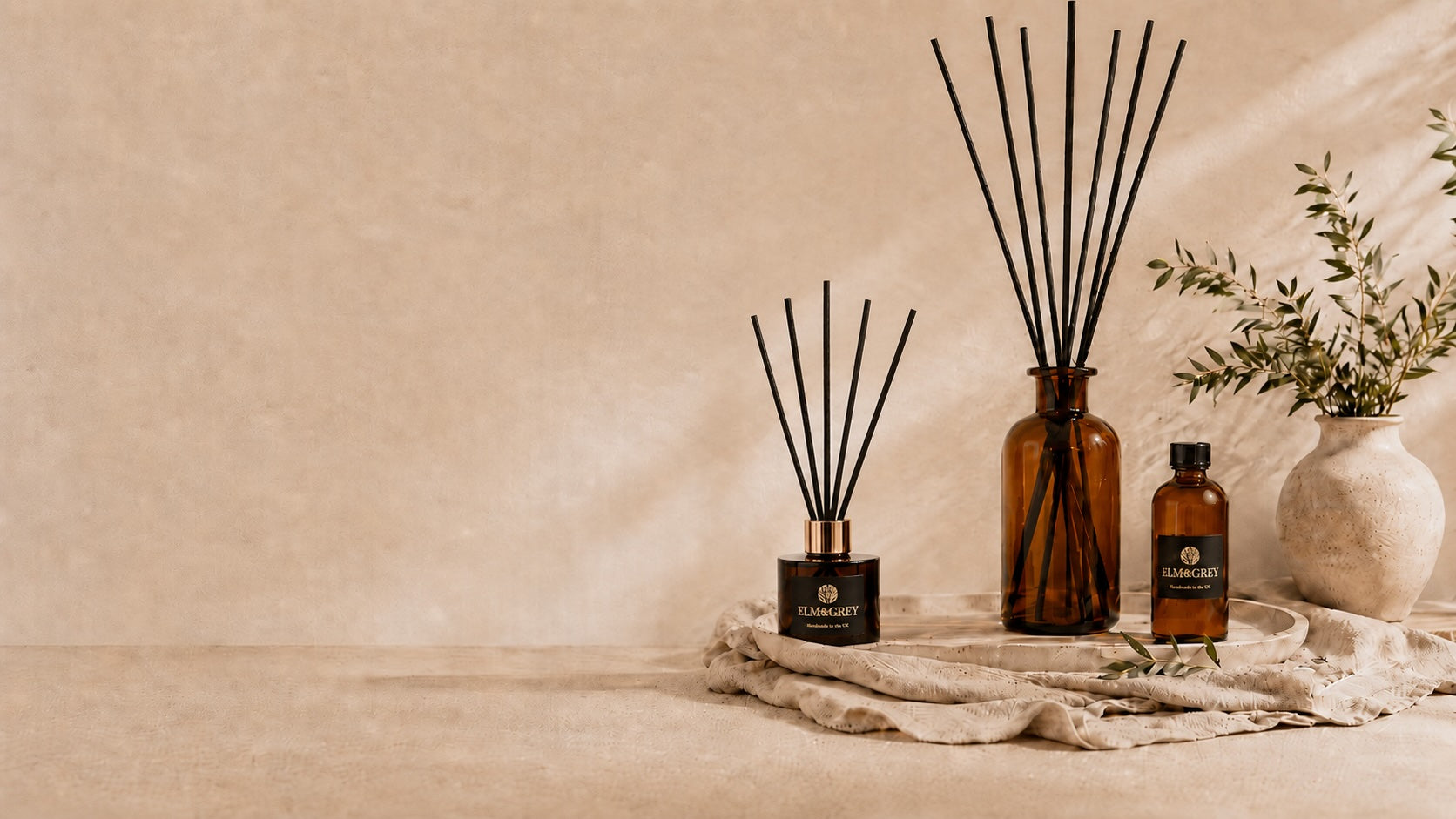 Elm & Grey reed diffuser and large amber glass statement diffuser with black reeds and refill bottle on linen cloth with olive branches